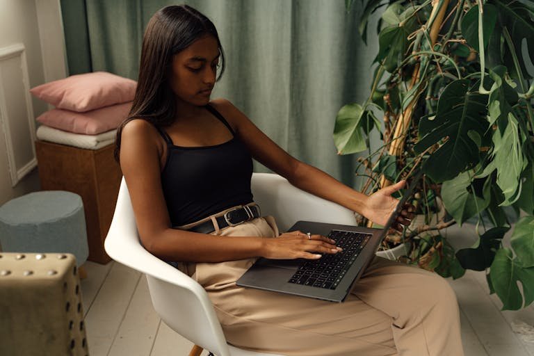 A young girl sits in a chair using a laptop, surrounded by plants in a cozy indoor setting.
